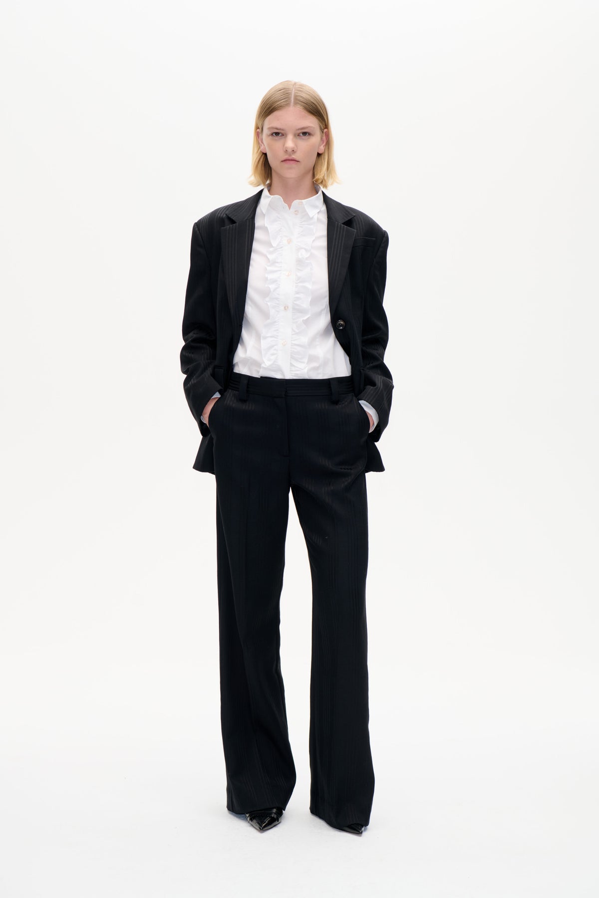 Frontal aufgenommenes Model in einem schwarzen Blazer mit Nadelstreifen und einer weitgeschnittenen Hose, dazu ein weißes Hemd mit Rüschen und Knopfleiste, vor schlichtem weißen Hintergrund.