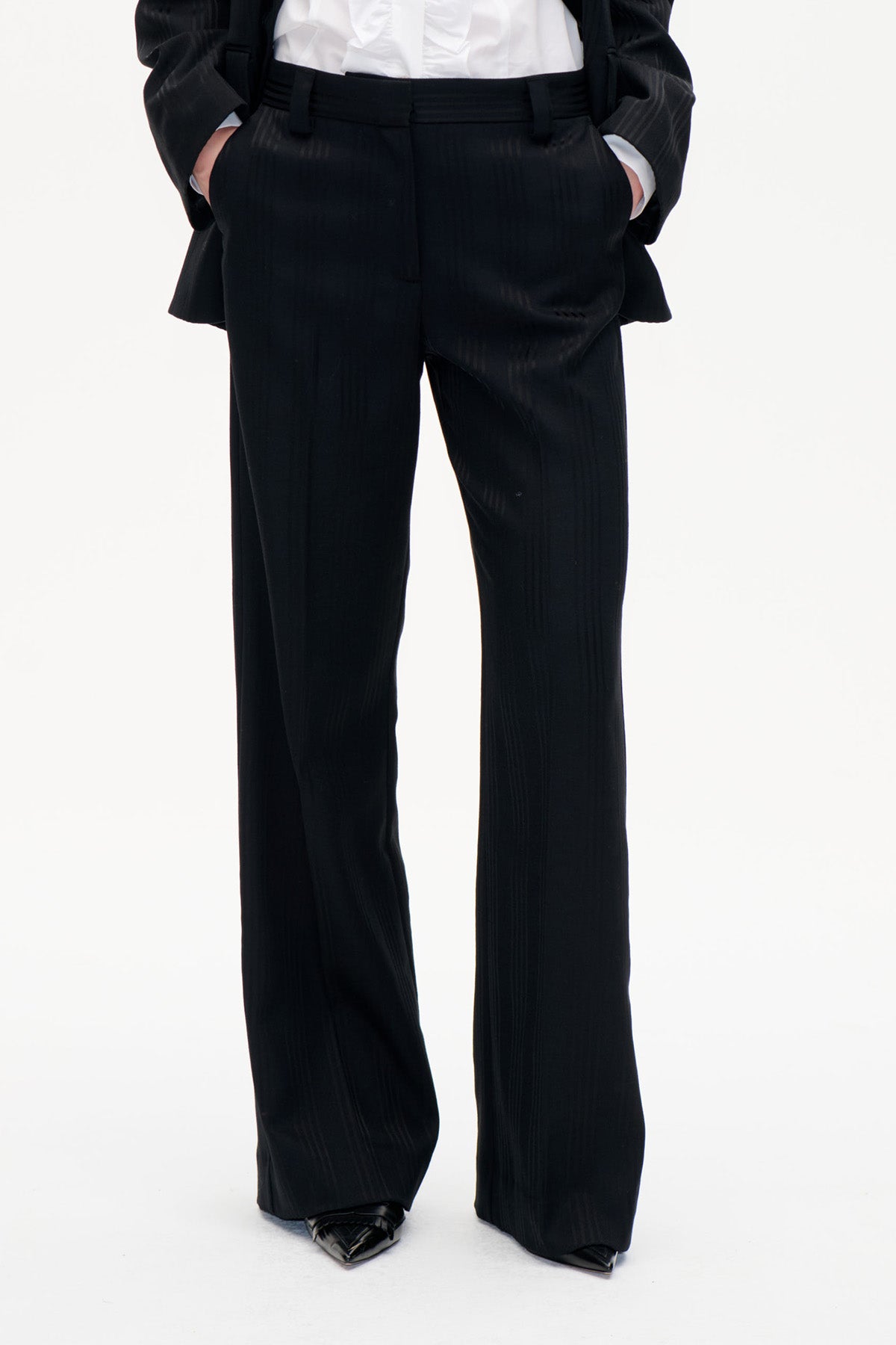 Vorderansicht einer schwarzen, weit geschnittenen Hose mit dezenten vertikalen Streifen und hohem Bund, kombiniert mit weißer Bluse und schwarzem Blazer.