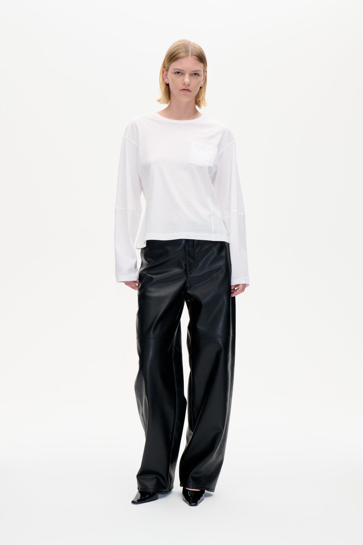 Model trägt ein weißes, langärmliges Rundhals-Top mit Brusttasche und eine schwarze, weit geschnittene Hose, von vorne gezeigt.