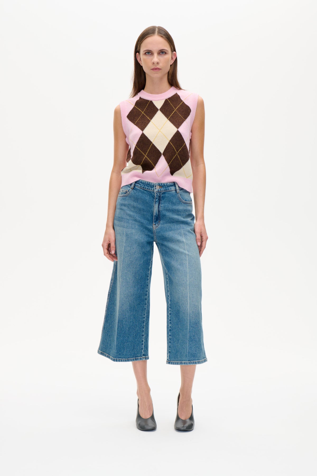 Vorderansicht eines Models, das eine ärmellose Strickweste mit Rautenmuster in Rosa, Creme und Braun sowie eine blaue Jeans mit weitem Bein trägt.