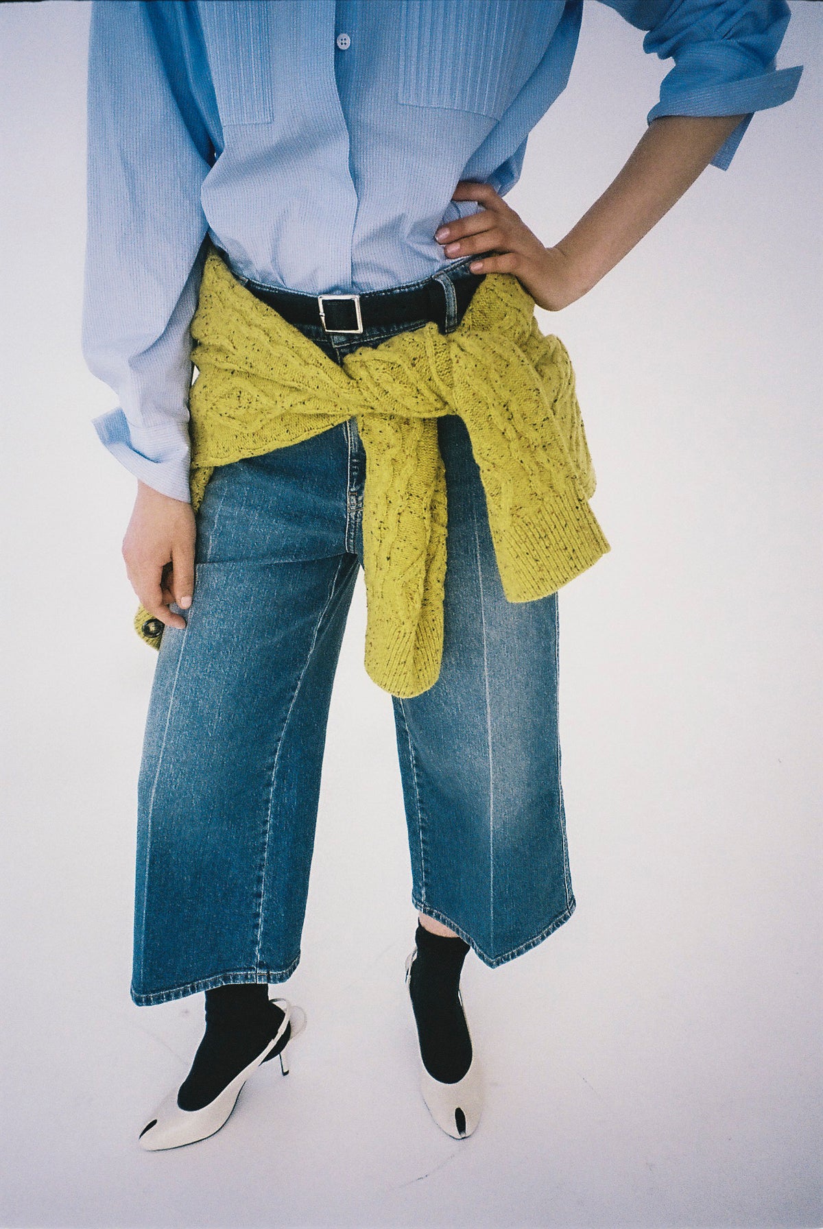 Gecropptes Kampagnenbild: Model trägt eine hellblaue Hemdbluse, weite blaue Jeans und einen senfgelben Strick, in der Taille geknotet.