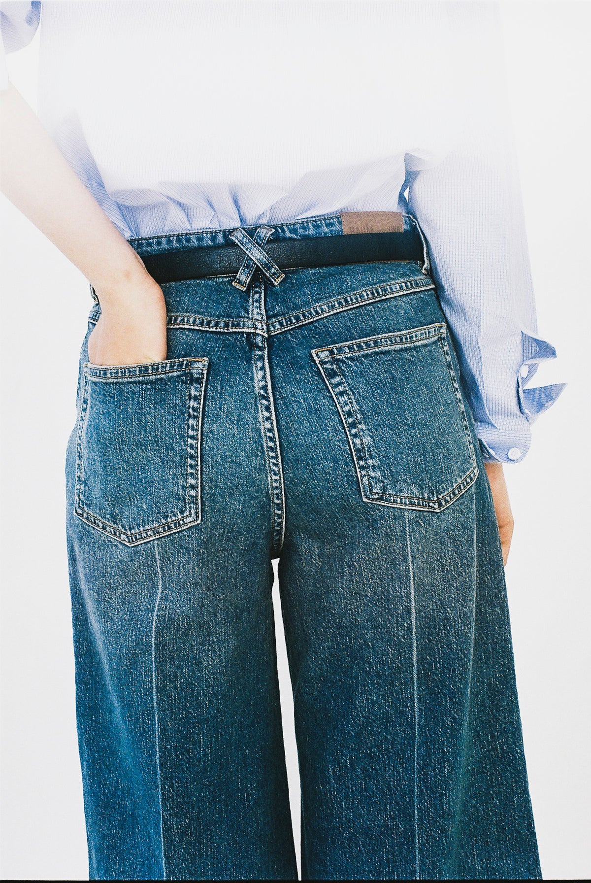 Nahaufnahme von hinten einer blauen Jeans mit weitem Bein, zwei aufgesetzten Taschen und einem hellblau gestreiften Hemd, das in den Bund gesteckt ist.
