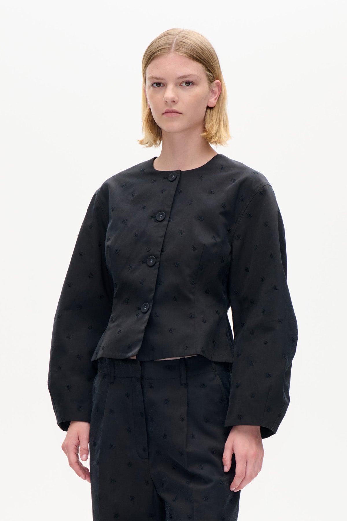 Model trägt eine schwarze, kurze Jacke mit dezenten Stickmotiven, Rundhalsausschnitt und großen Knöpfen, kombiniert mit passender Hose.