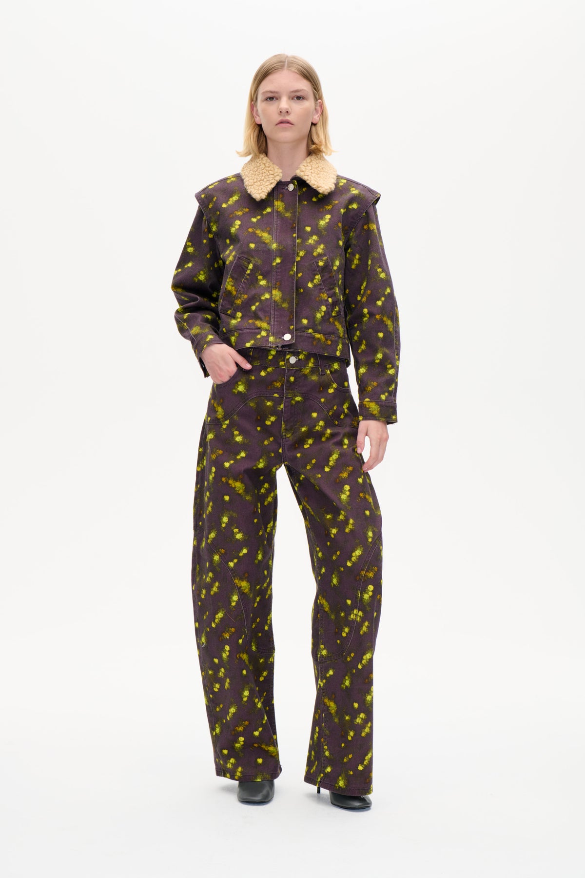 Vorderansicht eines Models, das eine anthrazit-violette Jacke mit leuchtend gelbem Sprenkelprint und cremefarbenem, strukturiertem Kragen trägt, dazu passende, weit geschnittene Hosen.