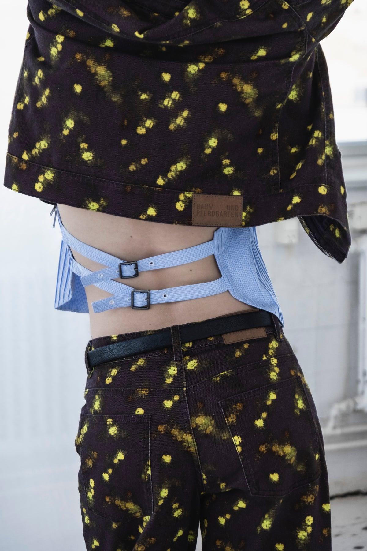Nahaufnahme des Kampagnenlooks: eine dunkelviolette Jacke mit Blumenmuster und gelb-grünen Blüten, darüber ein blau gestreiftes Cut-out-Top und eine dazu passende Hose.