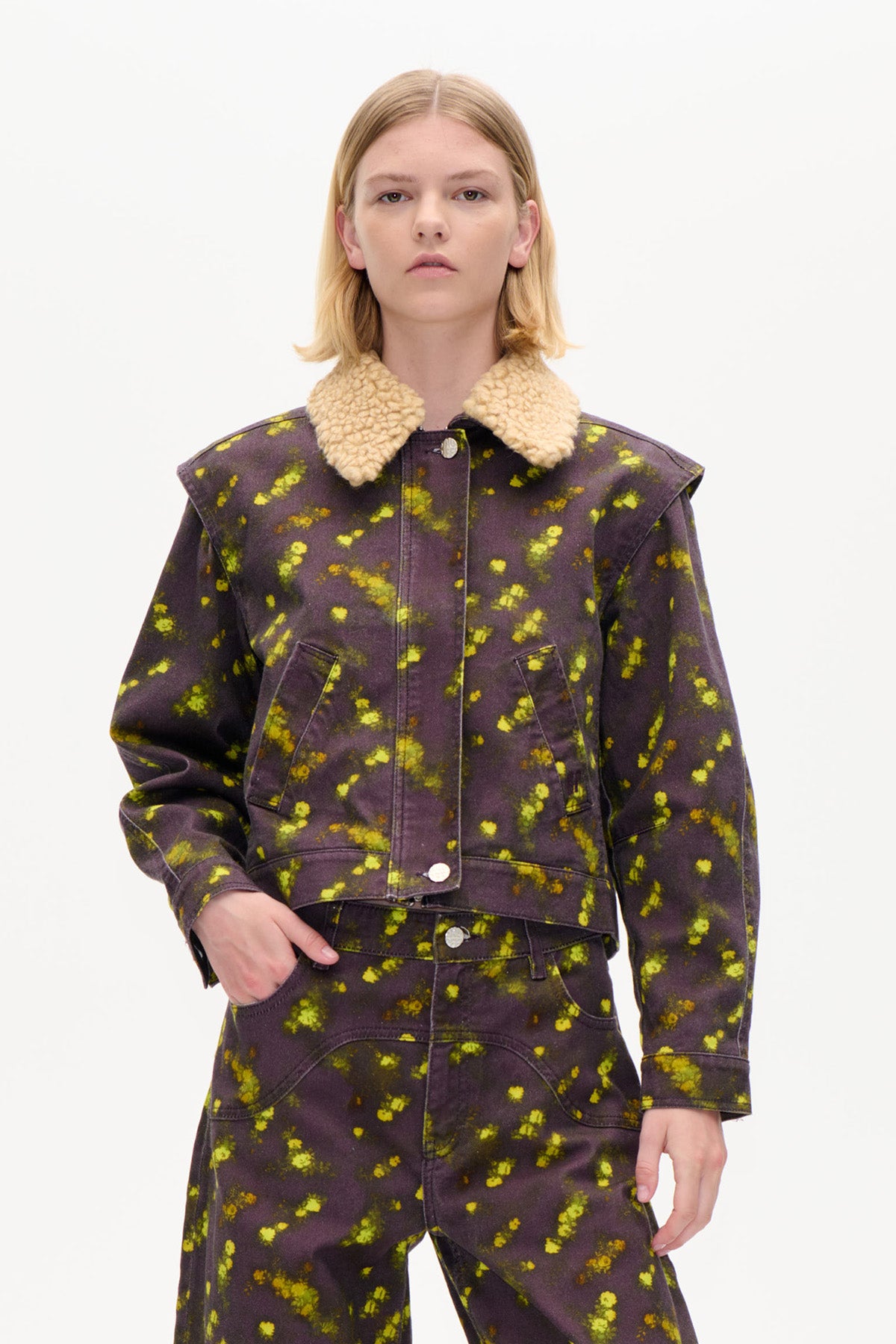 Vorderansicht einer kurz geschnittenen dunkelpflaumenfarbenen Jacke mit leuchtend gelbem, gesprenkeltem Muster, cremefarbenem Lammfellkragen und Druckknöpfen, getragen zu passenden Hosen.