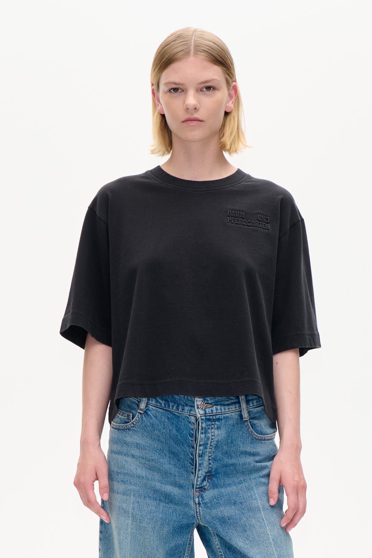 Vorderansicht eines schwarzen, verkürzten Oversized-T-Shirts mit kurzen Ärmeln und dezent erhabener Schrift auf der Brust, kombiniert mit blauen Jeans.