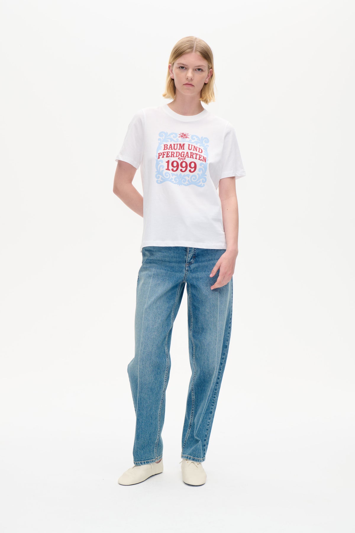 Model trägt ein weißes Kurzarm-T-Shirt mit blau-rotem Ornamentdruck „Baum und Pferdgarten 1999“ und blaue Jeans.