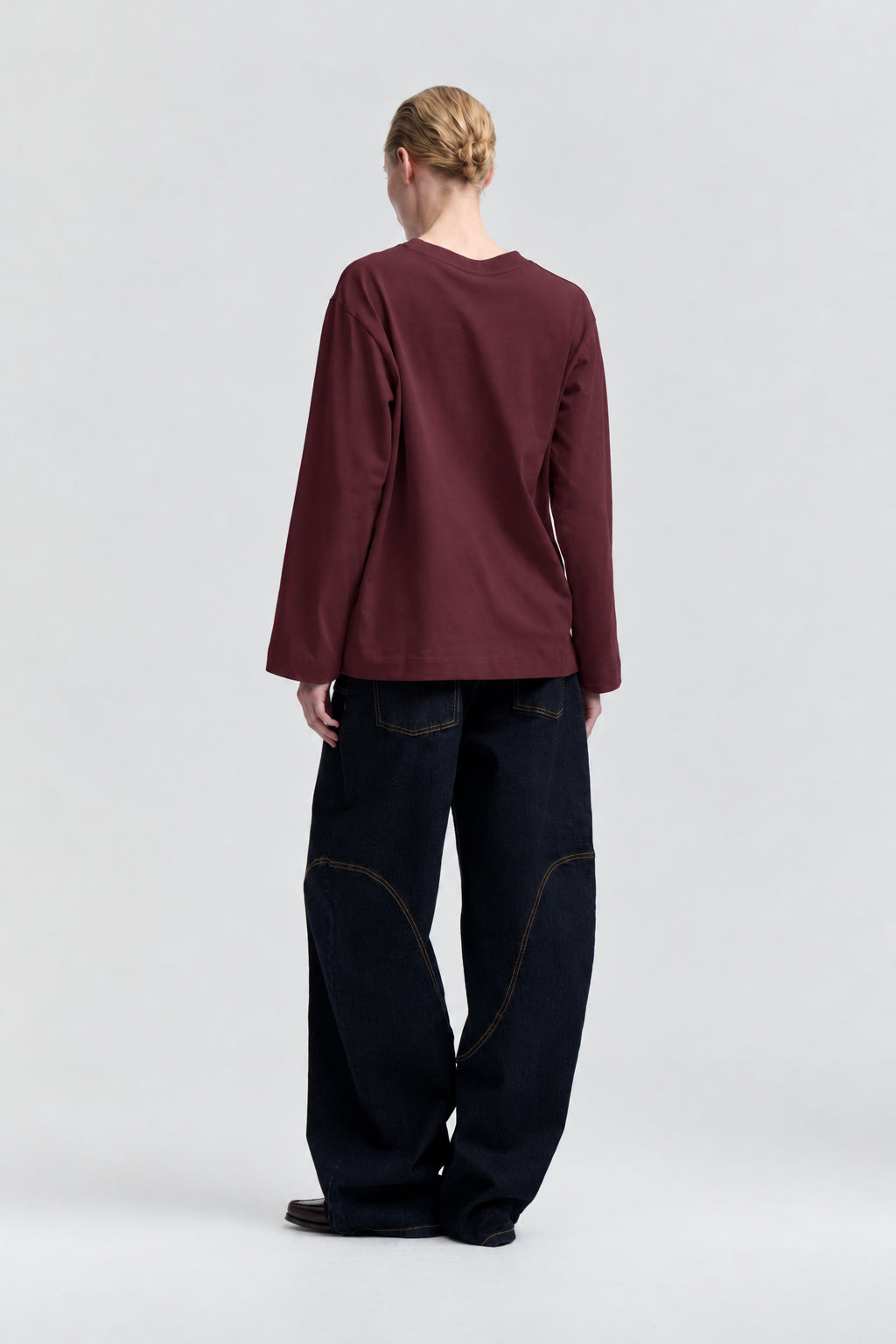 Rückenansicht eines Models in schlichtem burgunderrotem Langarmtop mit Rundhalsausschnitt und dunklen, weit geschnittenen Jeans.