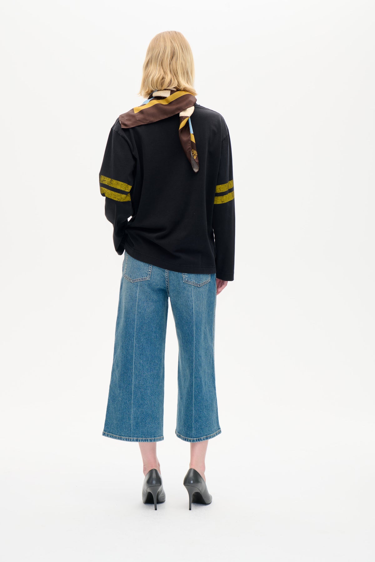 Rückenansicht eines Models, das ein schwarzes Langarmtop mit senffarbenen Ärmelstreifen, einen mehrfarbigen geometrischen Schal und weite blaue Jeans trägt.