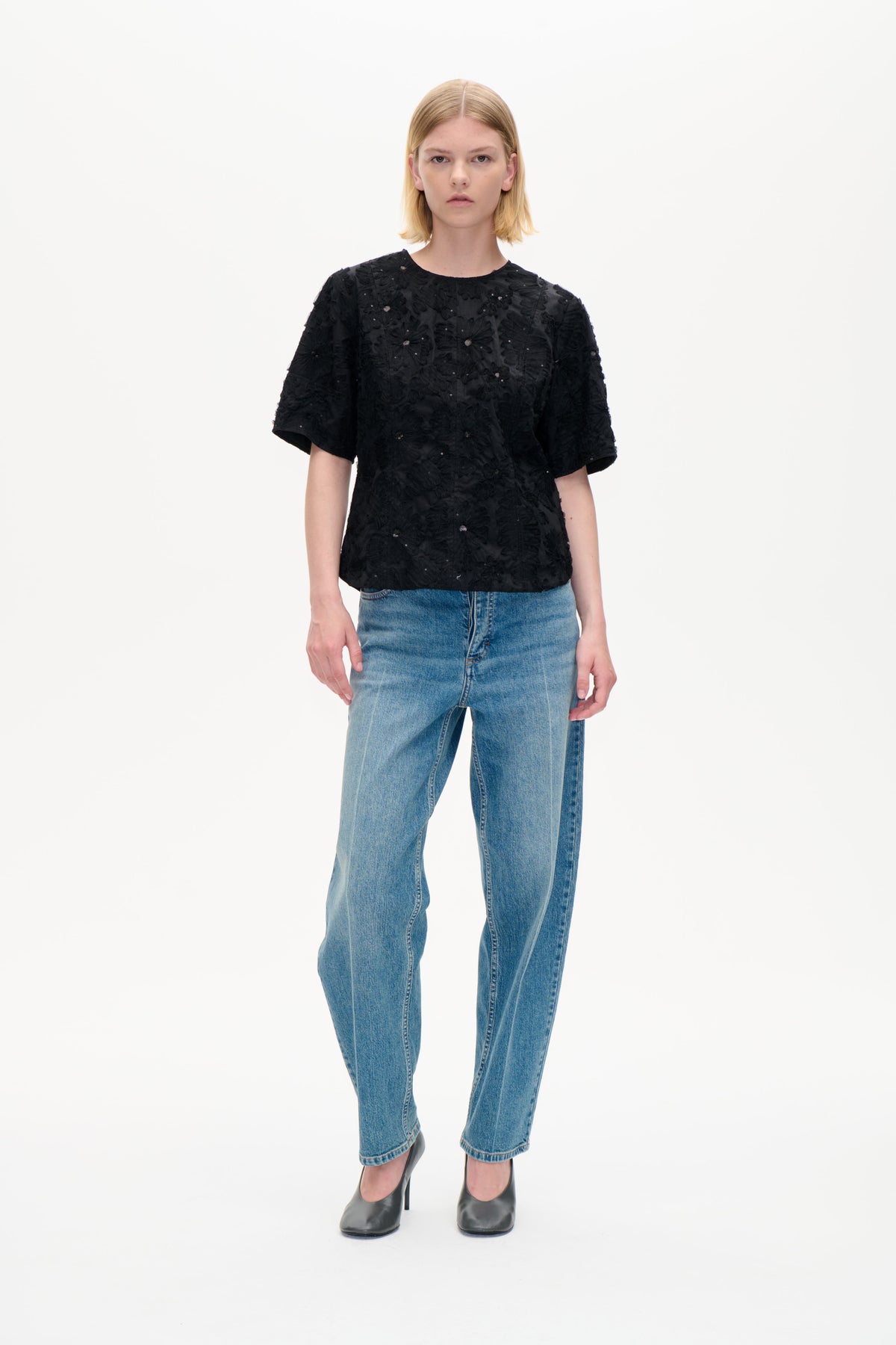 Vorderansicht eines Models, das ein schwarzes, kurzärmeliges Top mit erhabenen Blumenakzenten und kleinen Verzierungen zu blauen Jeans trägt.