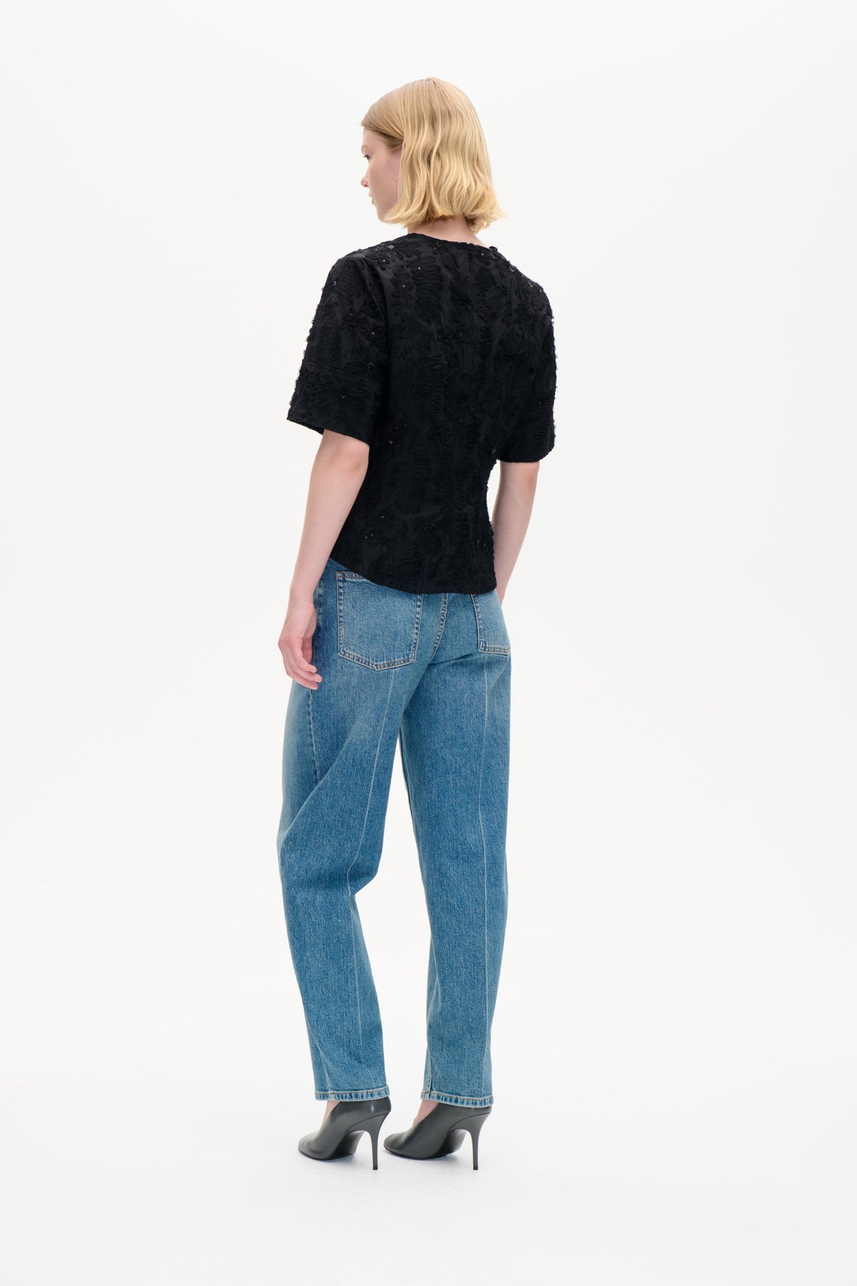 Model von hinten gesehen trägt ein schwarzes Kurzarm-Top mit strukturierten Blumenapplikationen und blauen Jeans.