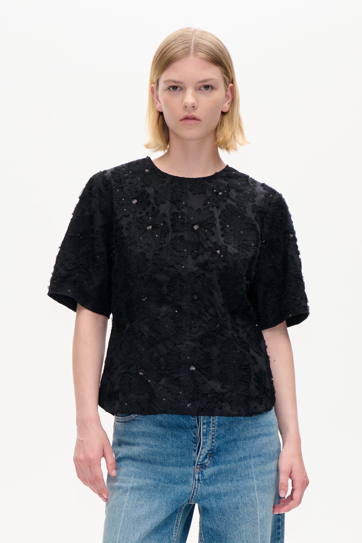 Vorderansicht eines schwarzen Kurzarmtops mit erhabener Blumenstickerei und kleinen Verzierungen, kombiniert mit blauer Jeans.