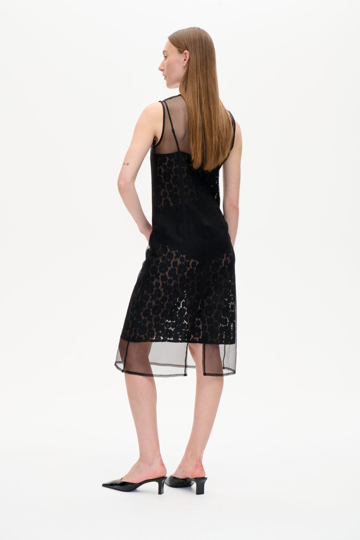 Rückenansicht eines Models, das ein schwarzes Spitzen-Slipkleid mit durchsichtigen Mesh-Einsätzen und transparentem, schwarz eingefasstem Saum trägt.
