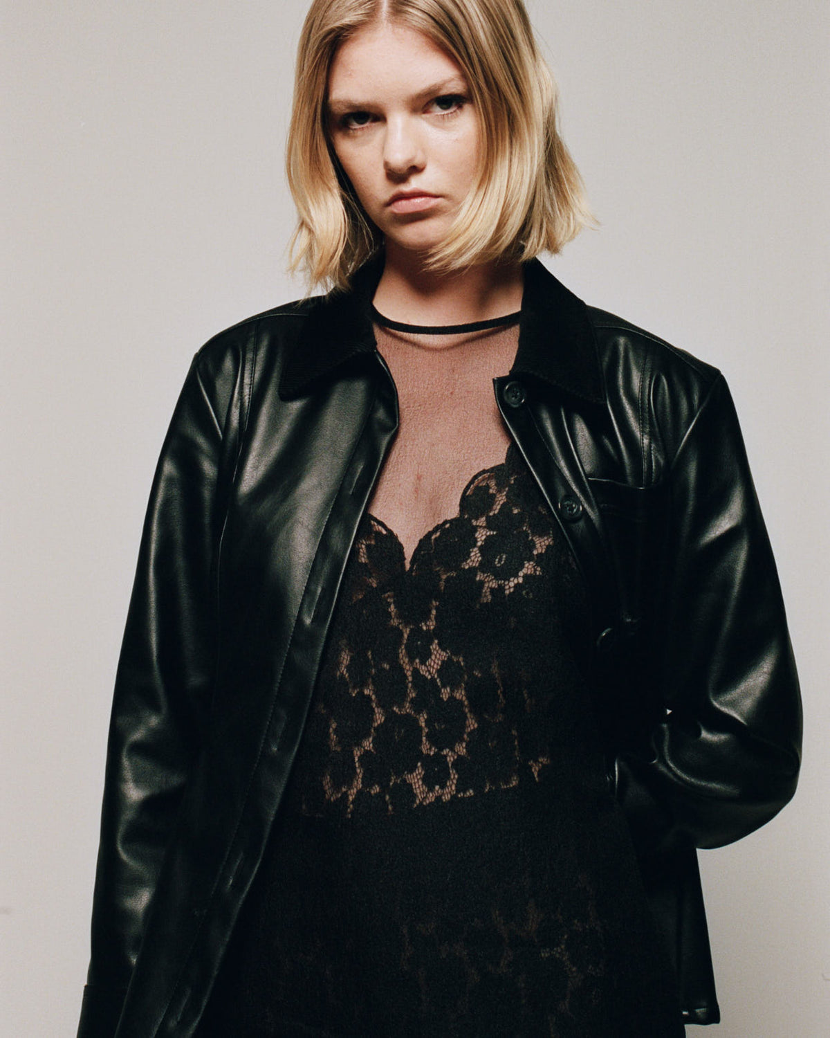 Kampagnenporträt eines Models, das eine glänzende schwarze Jacke mit Knopfleiste über einem schwarzen Spitzenkleid mit floralem Muster und transparentem roséfarbenen Ausschnitt trägt.