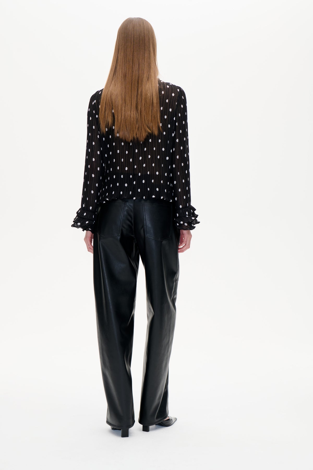 Rückenansicht einer schwarzen, transparenten, fein plissierten Bluse mit weißen Punkten und gerüschten Manschetten, kombiniert mit einer schwarzen Hose.