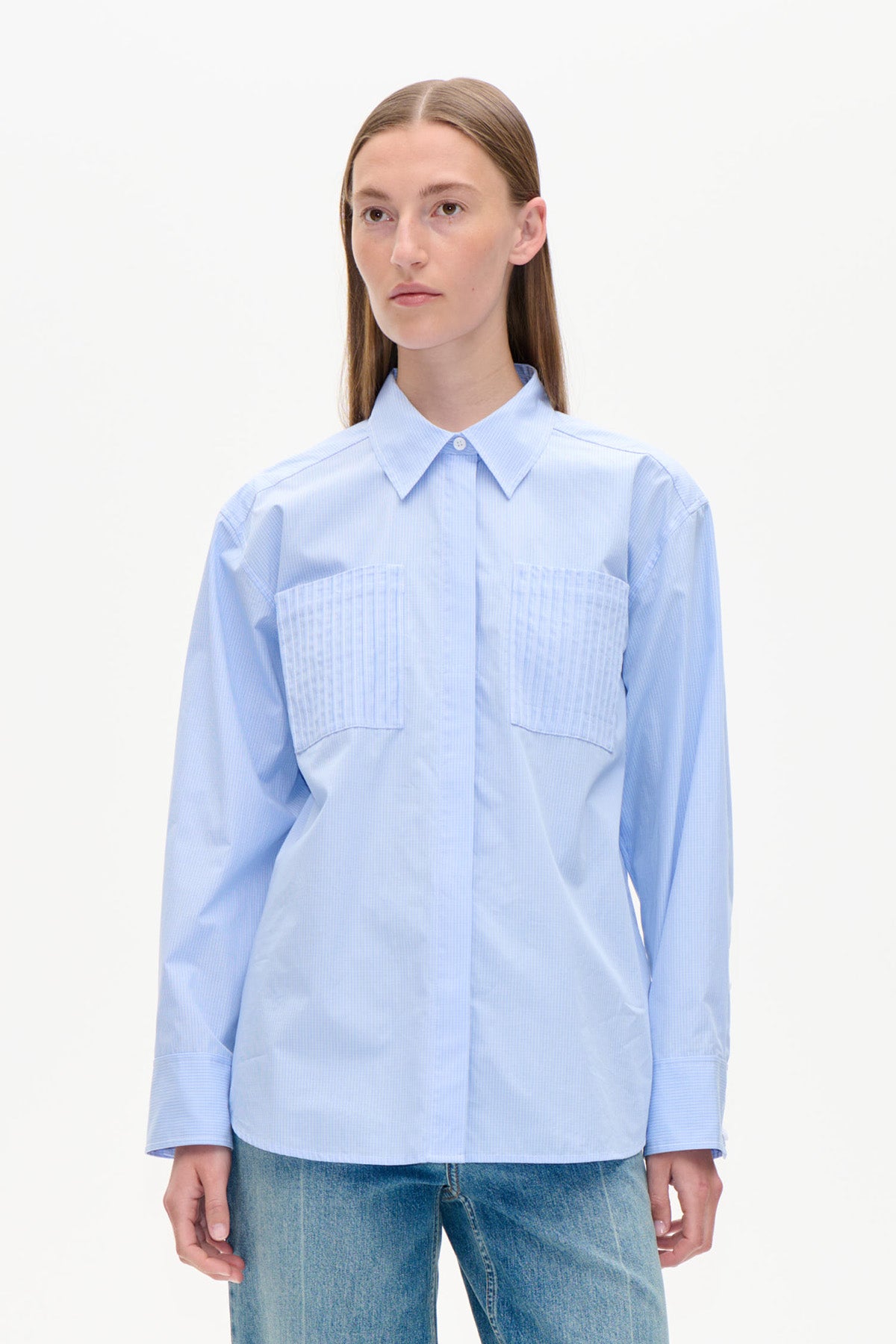 Vorderansicht einer hellblauen, langärmeligen Bluse mit spitzem Kragen, verdeckter Knopfleiste und zwei plissierten Brusttaschen.