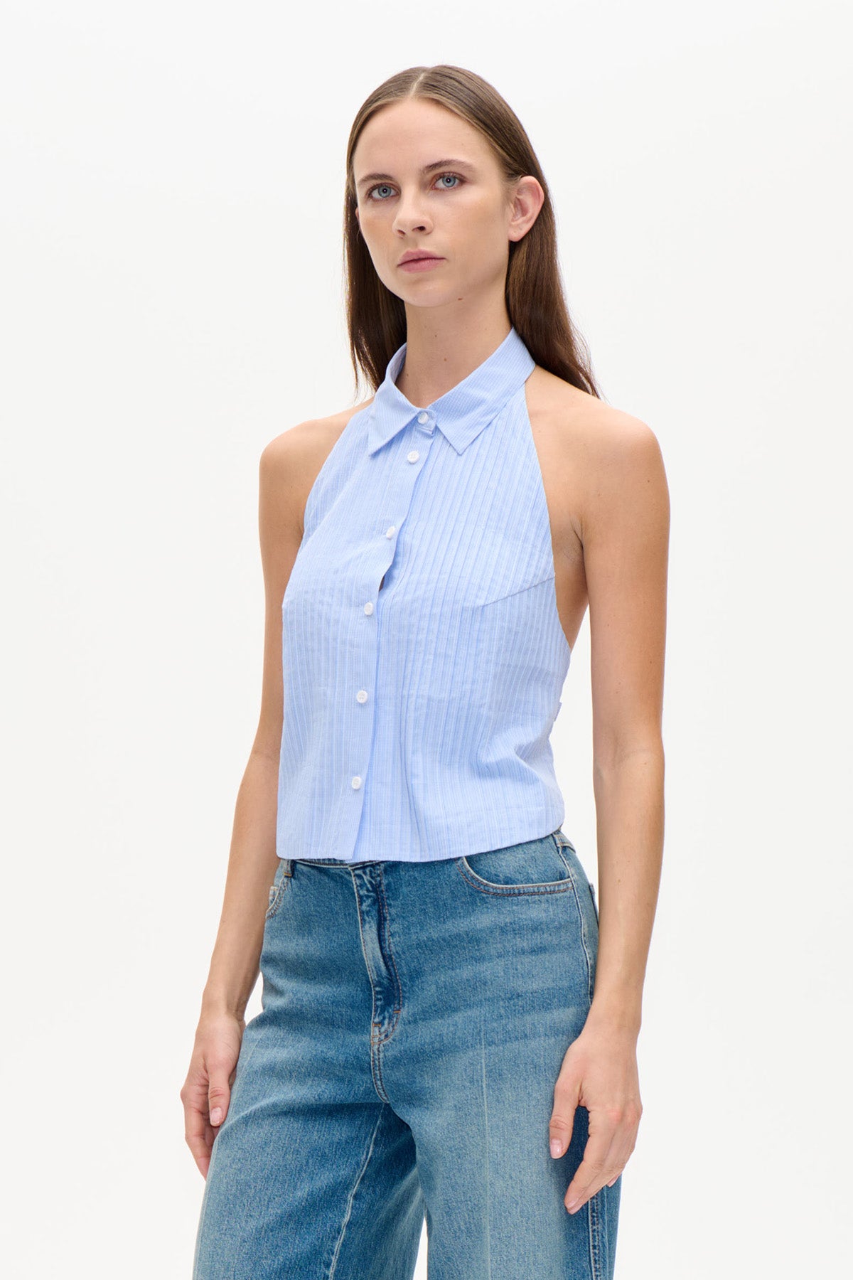 Model trägt ein hellblaues, fein gestreiftes, ärmelloses Oberteil mit Kragen, Knopfleiste und halterähnlichem Ausschnitt, dazu blaue Jeans.