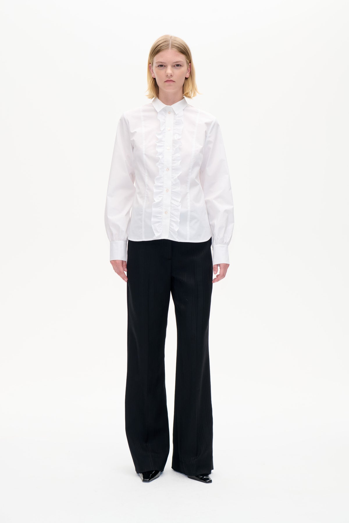 Frontal aufgenommenes Studio-Porträt eines Models, das eine weiße Bluse mit Rüschen vorne und Knopfleiste sowie eine schwarze, weit geschnittene Hose trägt.