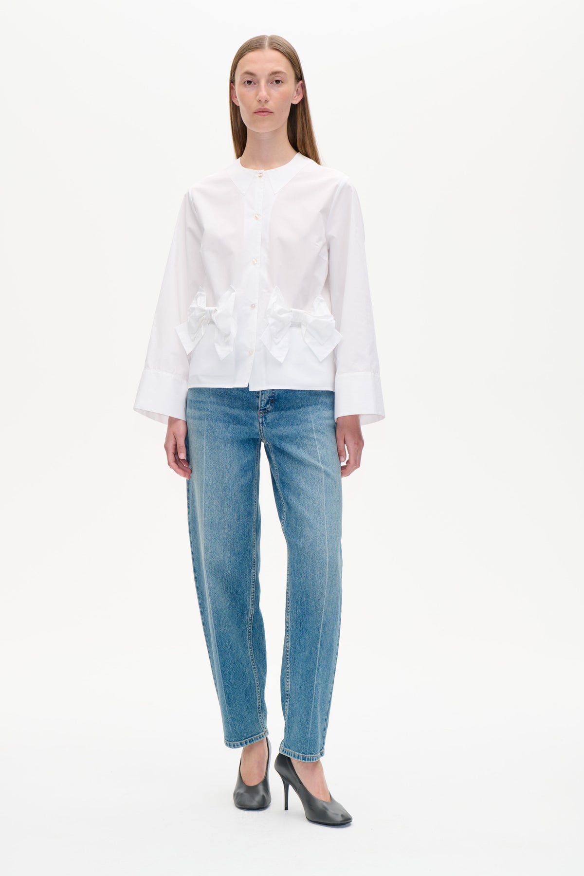 Frontansicht einer weißen Bluse mit Knopfleiste, übergroßem spitzem Kragen und skulpturalen Schleifendetails an der Taille, kombiniert mit blauen Jeans.