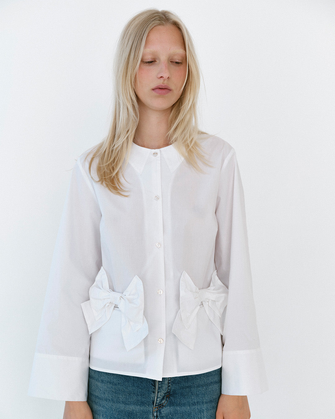 Model trägt eine weiße Bluse mit durchgehender Knopfleiste, spitzem Kragen, oversized Schleifen an der Taille und weiten Manschetten, kombiniert mit blauen Jeans.