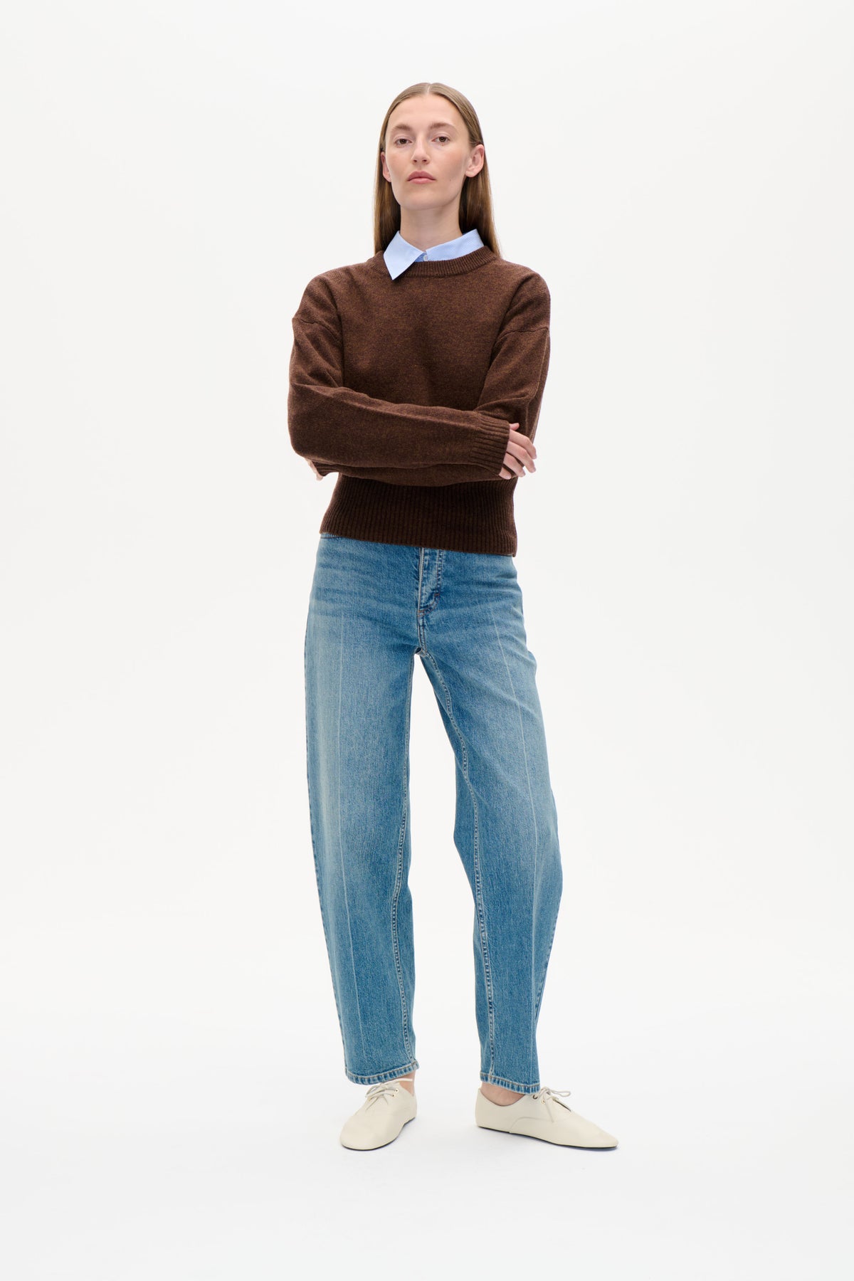 Vorderansicht eines Models, das einen braunen Rundhalspullover über einem hellblau gestreiften Hemd trägt, dazu blaue Jeans und cremefarbene Schnürschuhe.