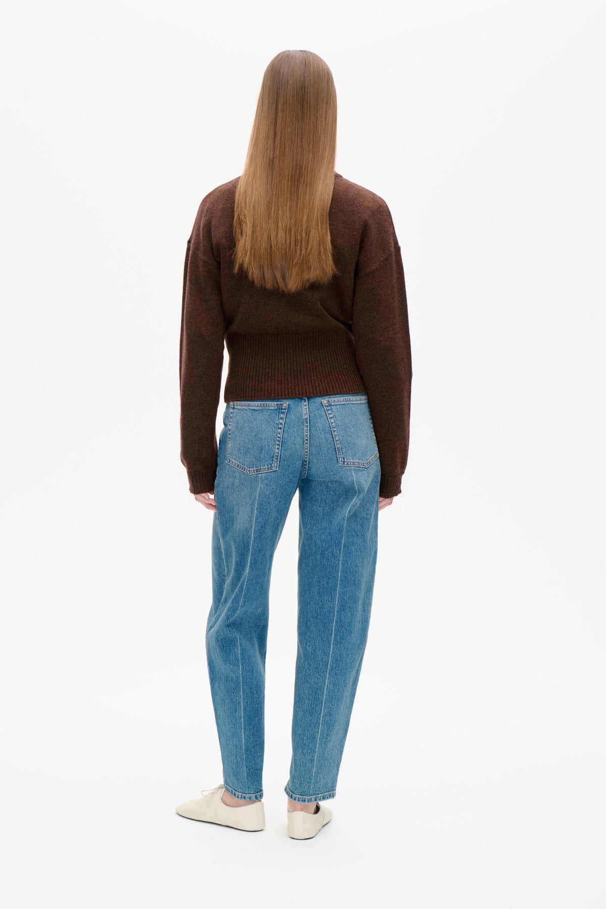 Rückansicht eines Models, das einen dunkelbraunen, langärmeligen Strickpullover mit geripptem Saum und eine blaue Jeans trägt.