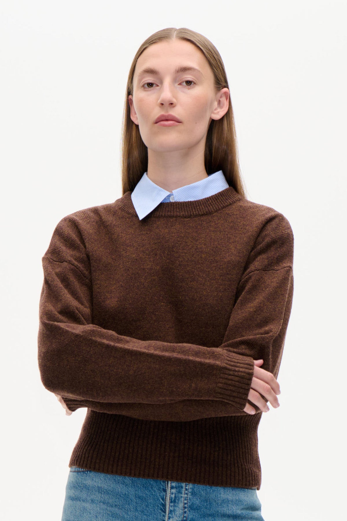 Vorderansicht eines Models, das einen braunen Rundhalspullover aus Strick über einem hellblauen Hemd mit Kragen trägt, kombiniert mit blauen Jeans.