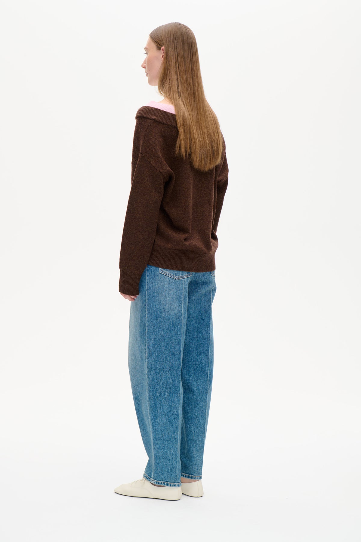 Rückenansicht eines Models in einem dunkelbraunen Pullover aus Strick mit weitem, umgeschlagenem Kragen, der eine hellrosa Lage darunter zeigt.
