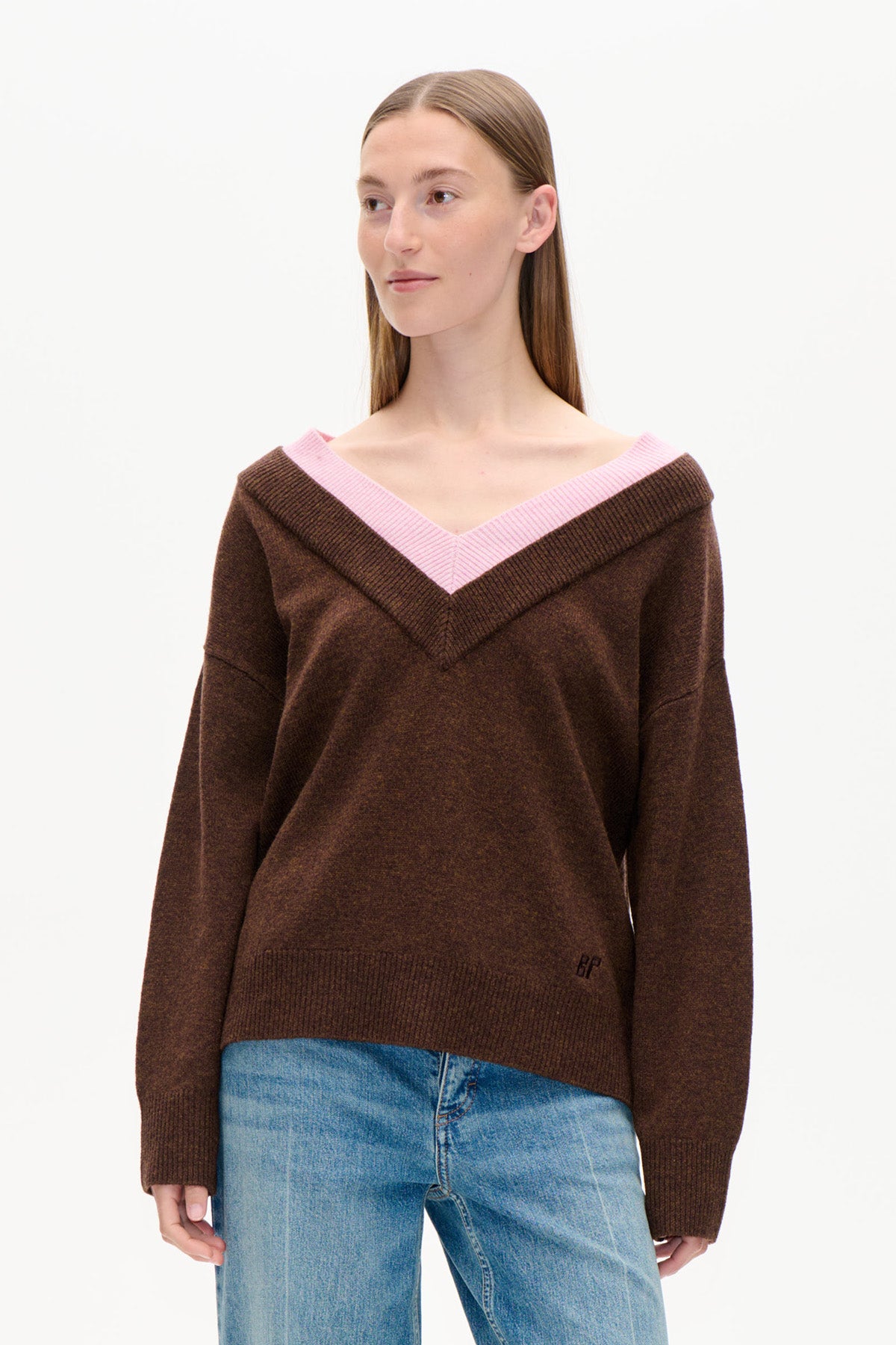 Vorderansicht eines dunkelbraunen Pullovers mit weitem, schulterfreiem V-Ausschnitt und rosa Rippenbündchen, kombiniert mit blauen Jeans.