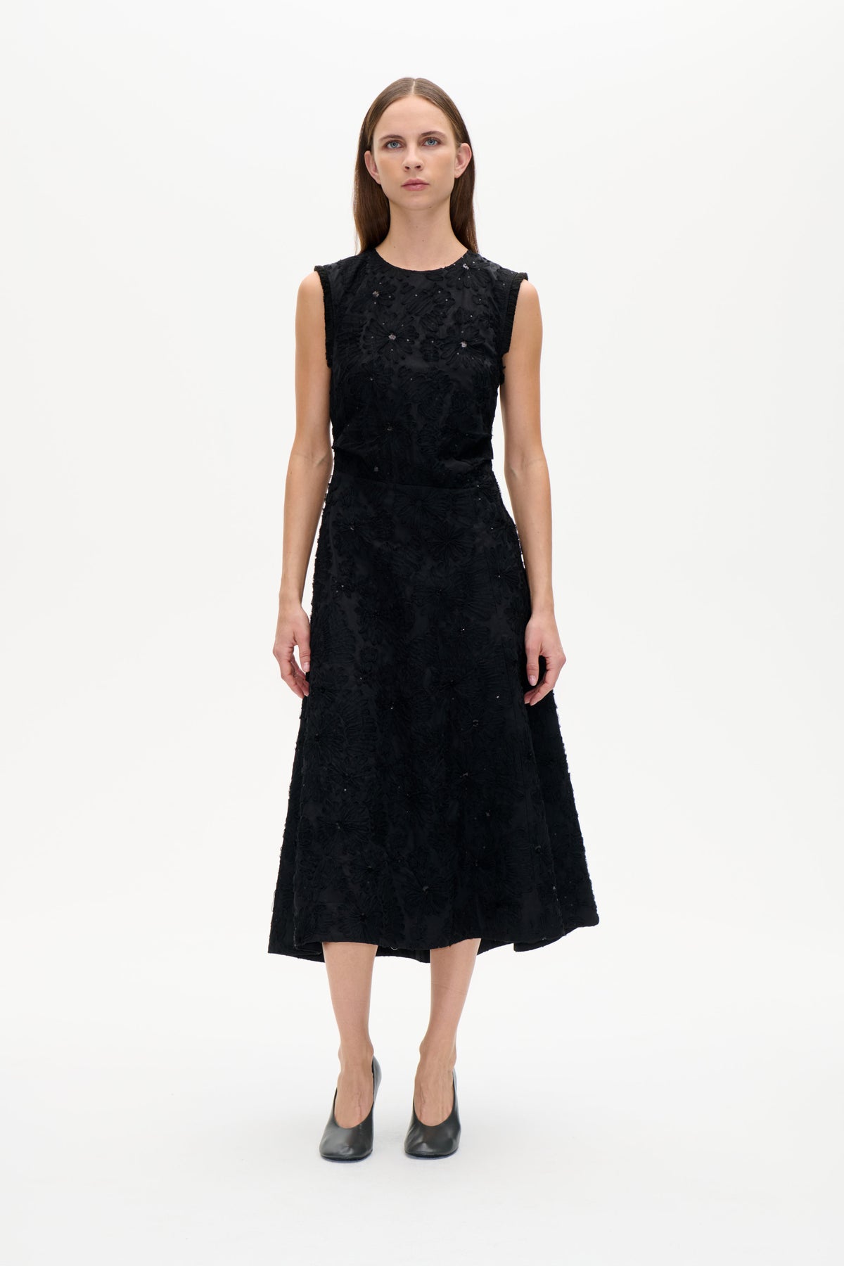 Vorderansicht eines Models in einem schwarzen ärmellosen Kleid mit erhabenen floralen Details und verstreuten Pailletten, tailliertem Schnitt und ausgestelltem Rock.