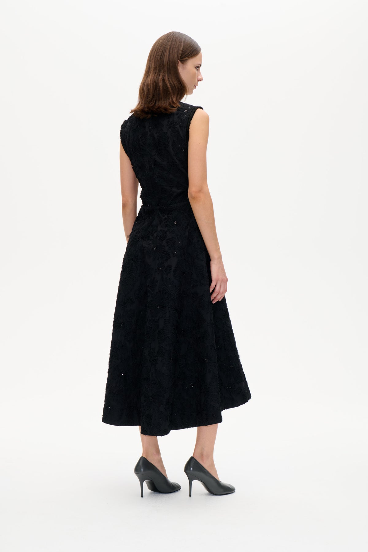 Model trägt ein schwarzes ärmelloses ausgestelltes Kleid mit erhabenen Blumendetails und verstreuten Verzierungen, gezeigt in seitlicher Rückansicht.