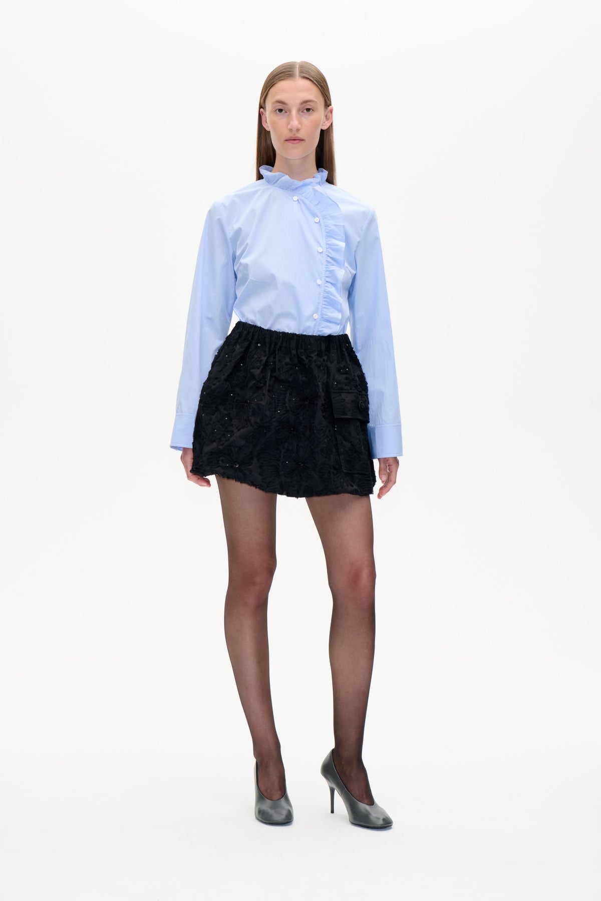 Modell von vorne in einer hellblauen, fein gestreiften Bluse mit Rüschenkragen und asymmetrischer Rüschenblende, kombiniert mit einem schwarzen, strukturierten Minirock.