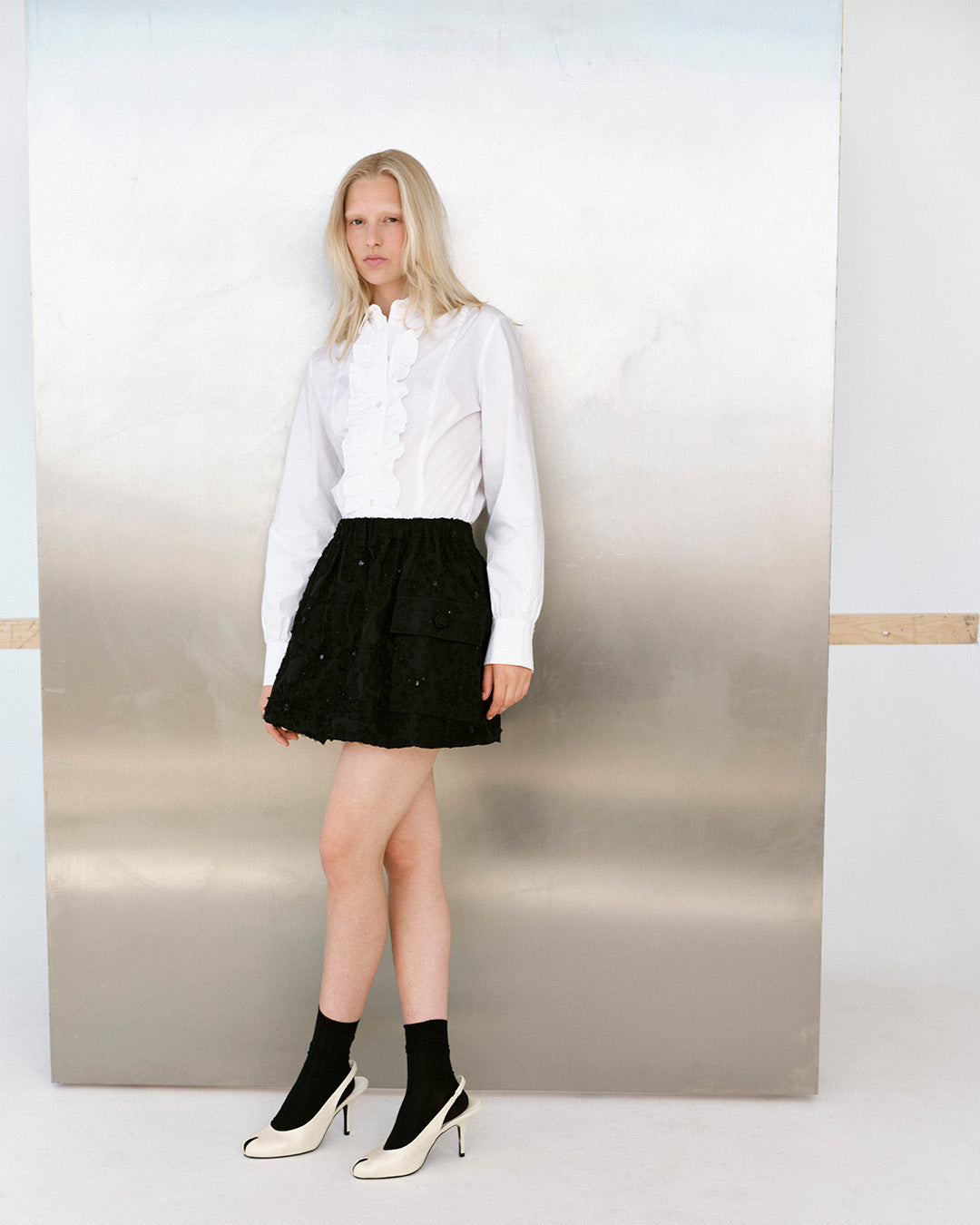 Kampagnenporträt eines Models, das eine weiße Rüschenbluse, einen schwarzen strukturierten Minirock, schwarze Socken und cremefarbene High Heels trägt.