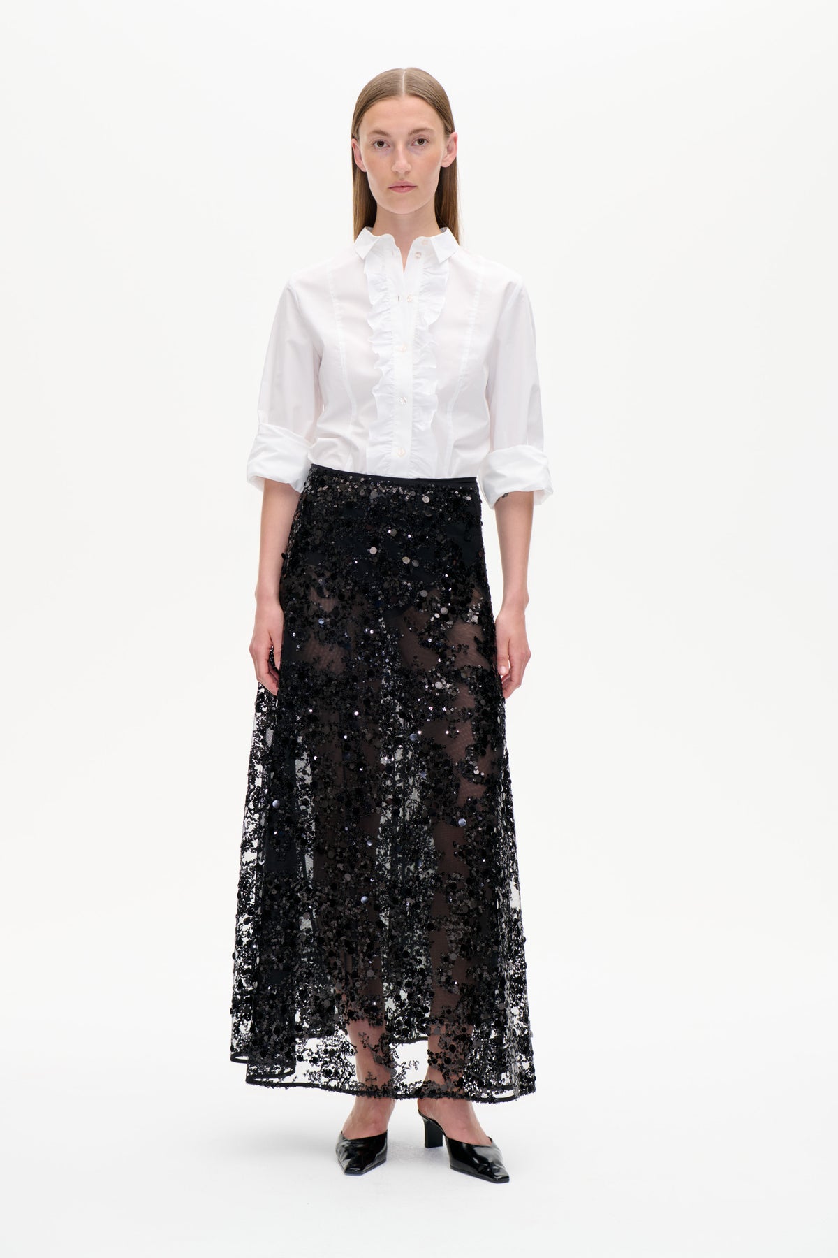 Vorderansicht eines Models, das eine weiße Bluse mit Rüschen und Knopfleiste sowie einen transparenten schwarzen Pailletten-Midi-Rock vor weißem Hintergrund trägt.