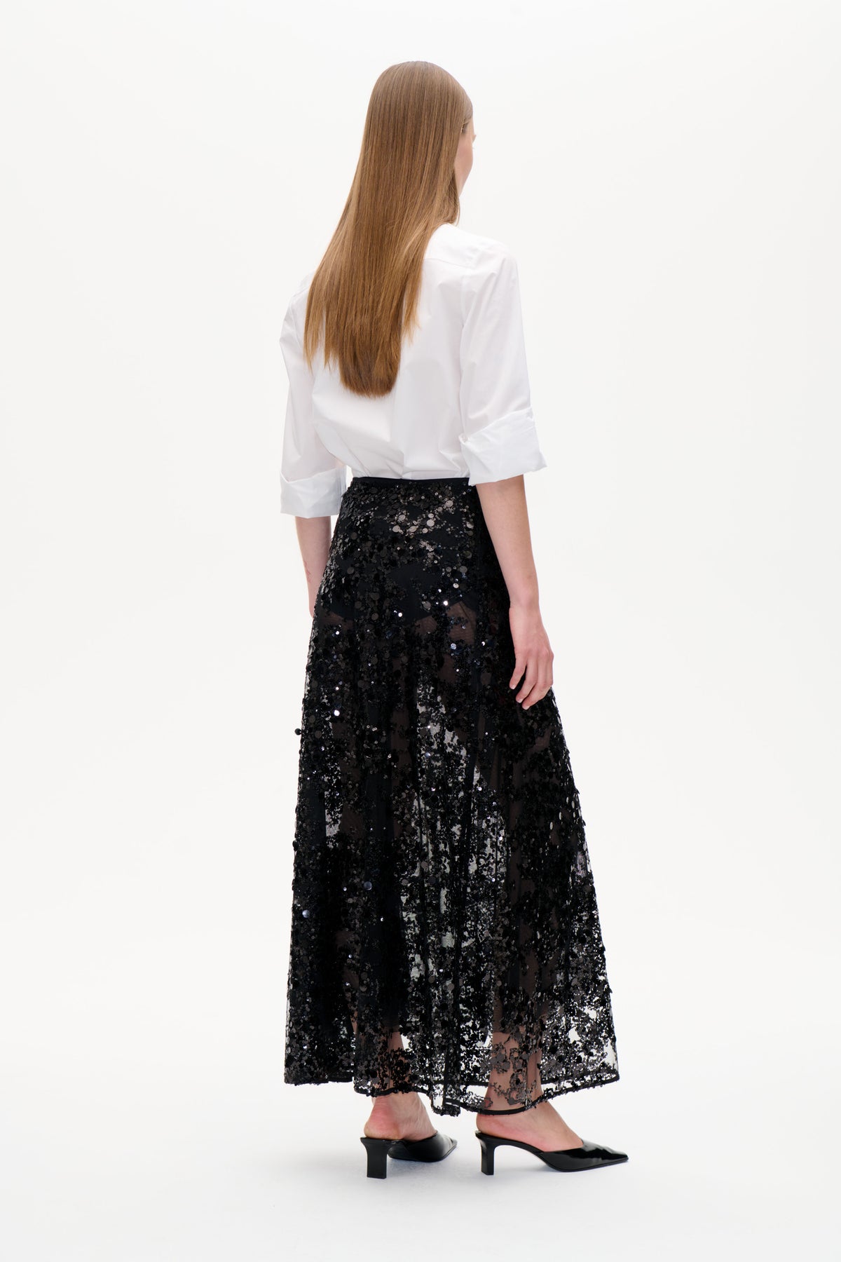 Rückenansicht eines Models, das eine weiße Bluse und einen schwarzen, transparenten midi-Rock trägt, der dicht mit Pailletten besetzt ist.