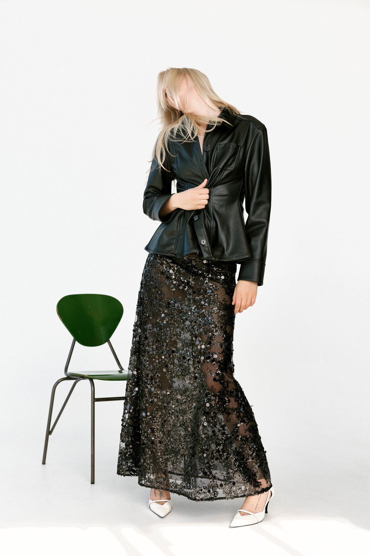 Kampagnenporträt eines Models in einer schwarzen Jacke mit Knopfleiste und einem halbtransparenten, schwarz-paillettenbesetzten Midirock, neben einem grünen Stuhl vor weißem Hintergrund.