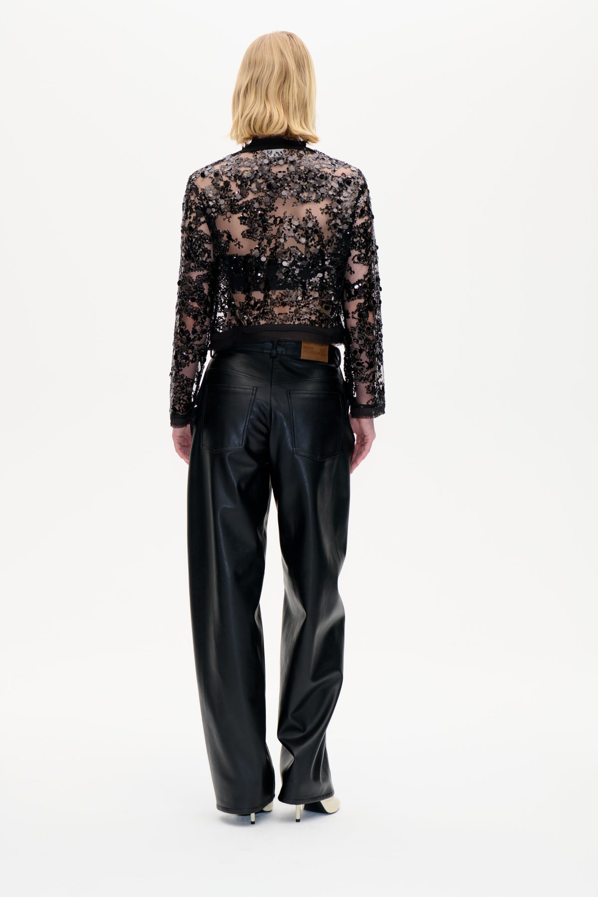 Rückenansicht eines Models, das ein durchsichtiges schwarzes Langarmtop mit Pailletten und eine weite schwarze Hose trägt.