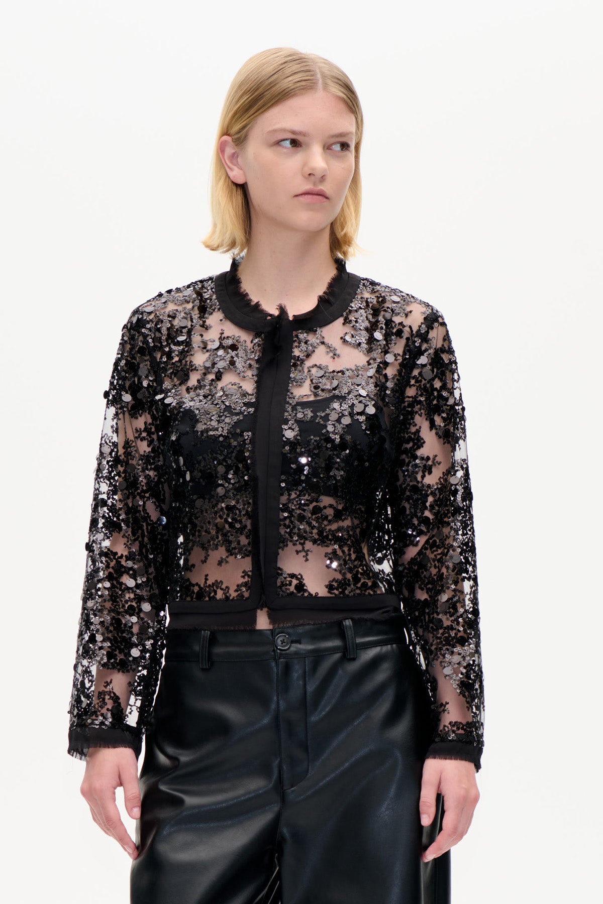 Frontalansicht einer transparenten schwarzen Langarmbluse mit Pailletten, schwarzem Kragenbesatz und knopflosem Vorderteil, kombiniert mit einer schwarzen Hose.