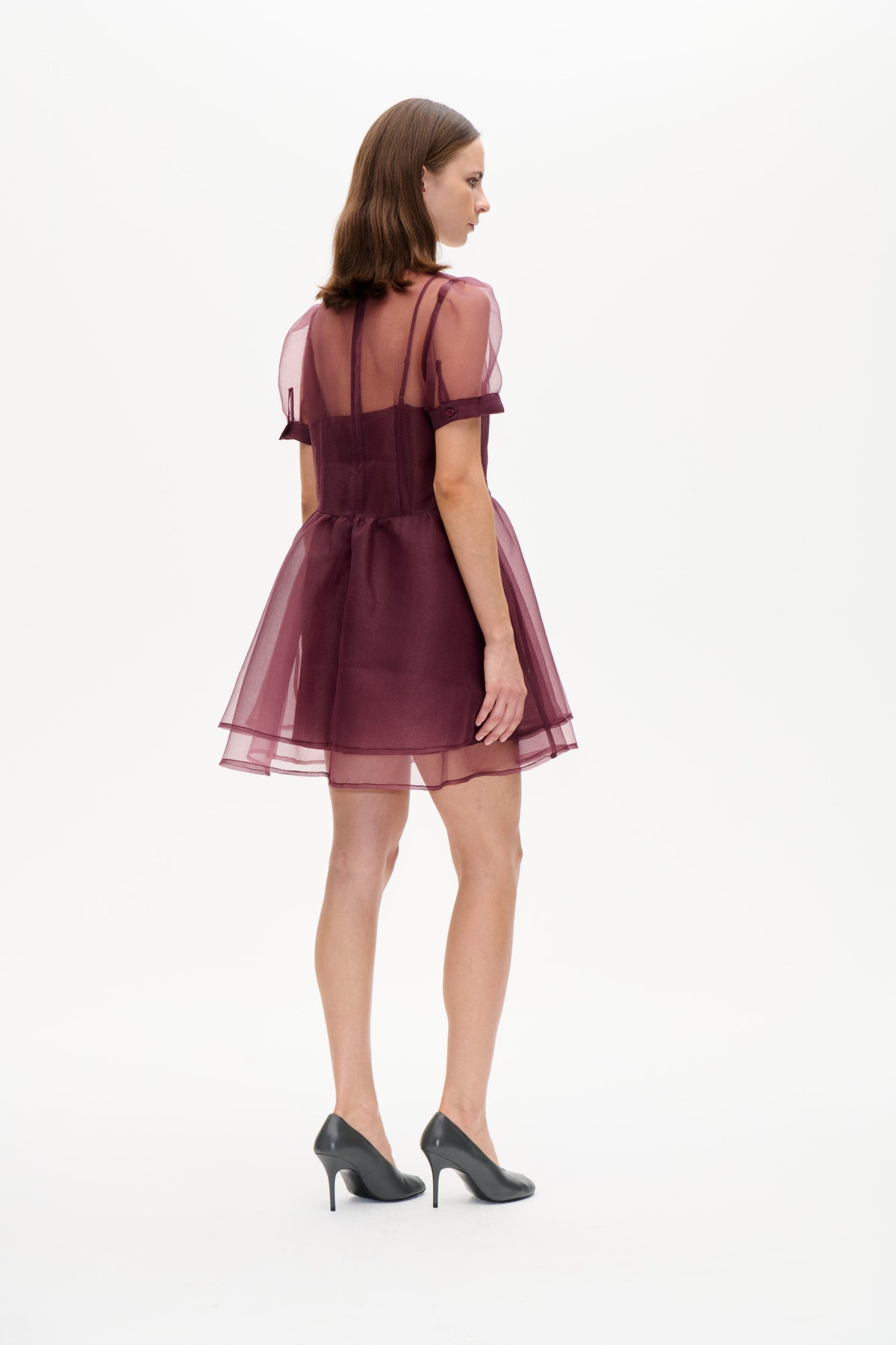 Model in einem pflaumenfarbenen Minikleid mit transparenten, mauvefarbenen Puffärmeln und einem gestuften, durchscheinenden Rock, von hinten in Dreiviertelansicht gezeigt.