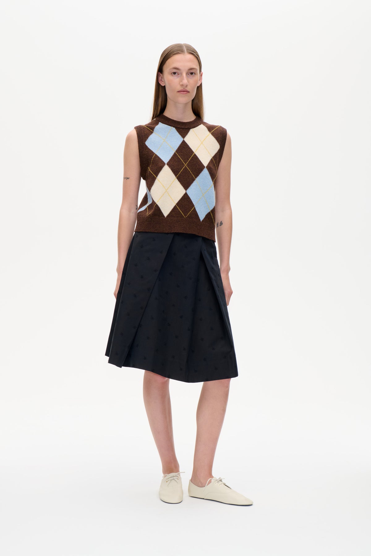Ein Model vor weißem Hintergrund trägt eine braune, ärmellose Strickweste mit blau- und cremefarbenen Rauten im Argyle‑Stil und einen schwarzen, gemusterten Rock.