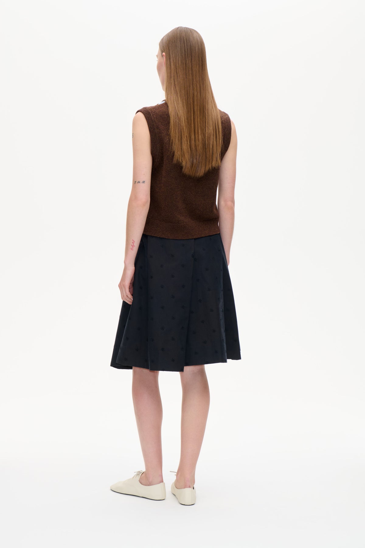Rückenansicht eines Models, das ein dunkelbraunes, ärmelloses Stricktop, einen schwarzen, gemusterten Midirock und cremefarbene flache Schuhe trägt.