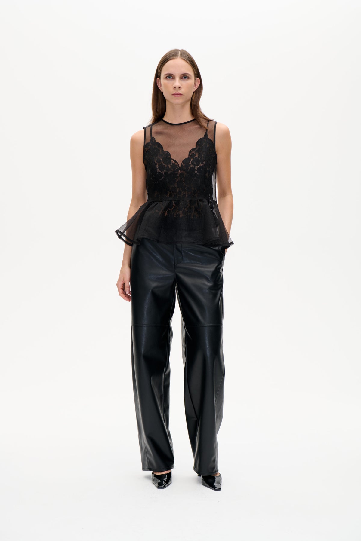 Model trägt ein schwarzes, transparentes, ärmelloses Top mit floralen Spitzeneinsätzen und ausgestelltem Saum, kombiniert mit glänzenden, weit geschnittenen Hosen.