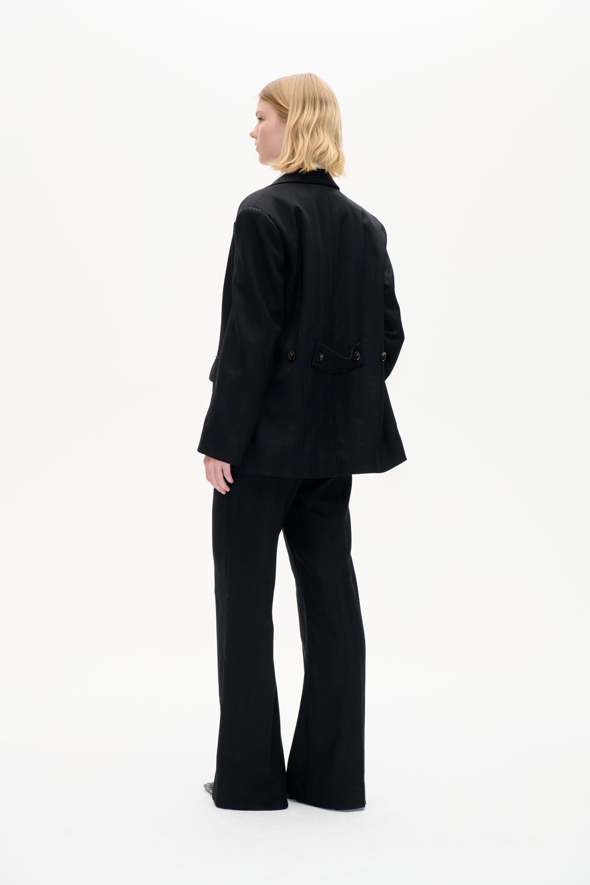 Model in einem schwarzen Nadelstreifenblazer mit einem am Rücken geknöpften Taillenriegel und einer Hose mit weitem Bein, seitlich und von hinten gezeigt.