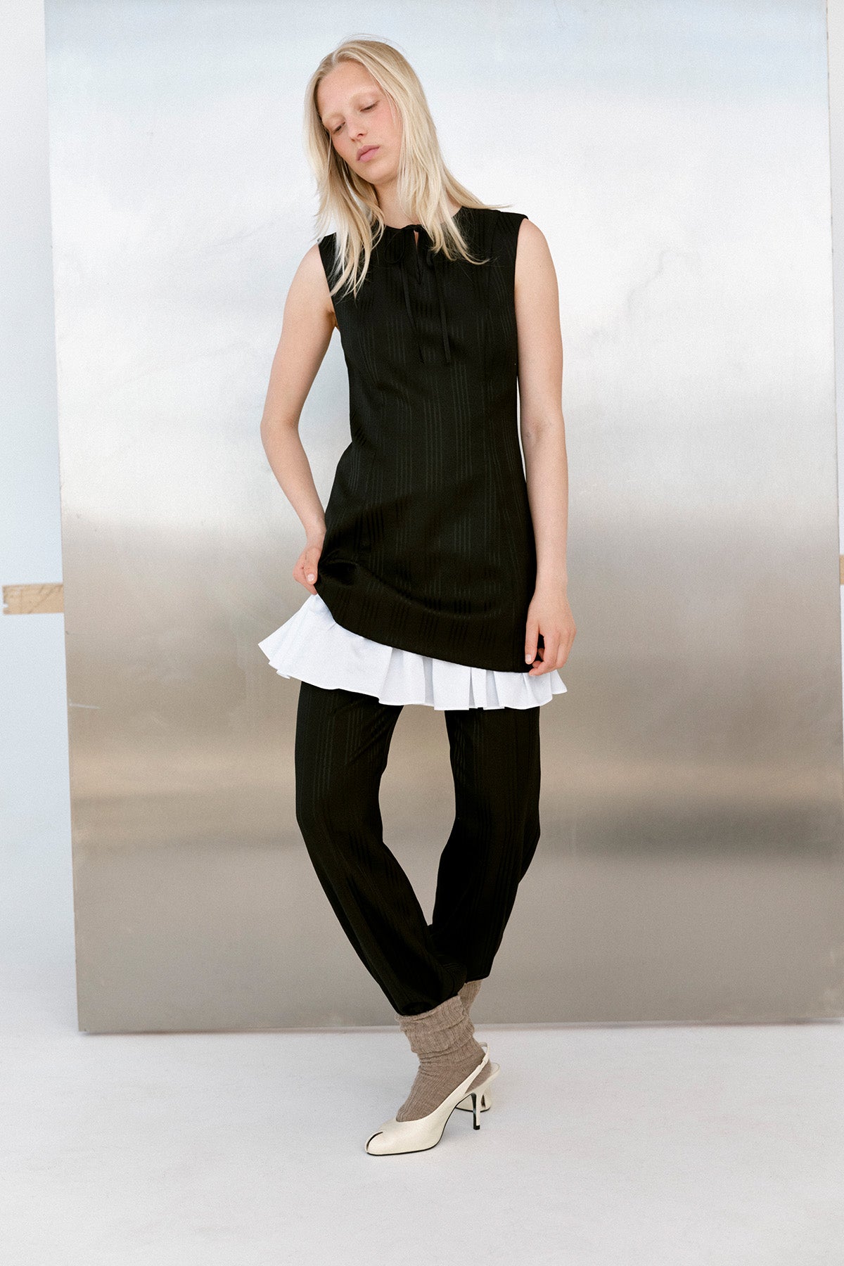 Model trägt eine ärmellose schwarze Tunika mit dezenten vertikalen Streifen und Bindeband am Ausschnitt, kombiniert mit einem weißen Top mit Rüschensaum und passenden Hosen.