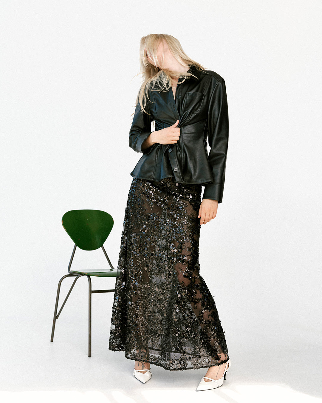 Model trägt eine glänzende schwarze Jacke mit durchgehender Knopfleiste sowie einen transparenten schwarzen Paillettenmaxirock und steht neben einem grünen Stuhl in weißen Highheels.