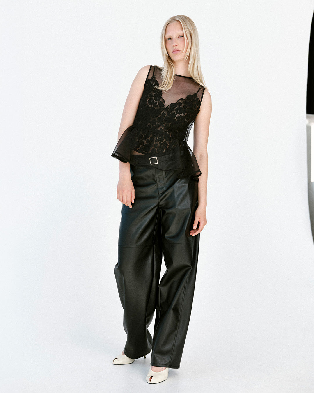 Model trägt ein transparentes schwarzes Spitzen-Peplum-Top, eine weite Hose im Lederlook und weiße Schuhe vor weißem Hintergrund.