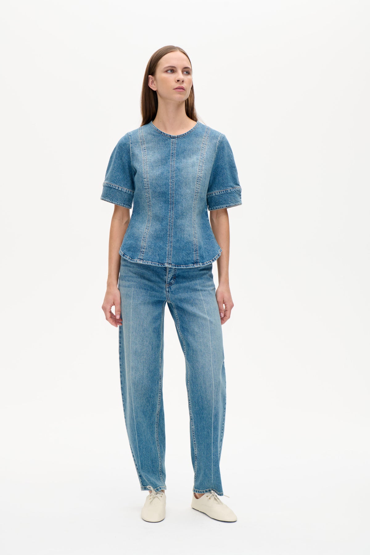 Vorderansicht eines Models in blauem Denim-Kurzarmtop mit rundem Halsausschnitt und markanten vertikalen Teilungsnähten, kombiniert mit Jeans.