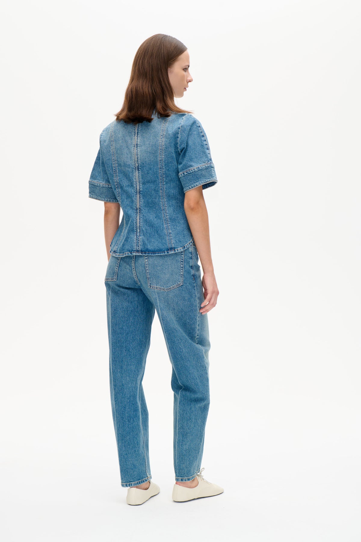 Model trägt ein blaues denim-Top mit kurzen Ärmeln und eine passende Jeans, von hinten und seitlich vor weißem Hintergrund fotografiert.