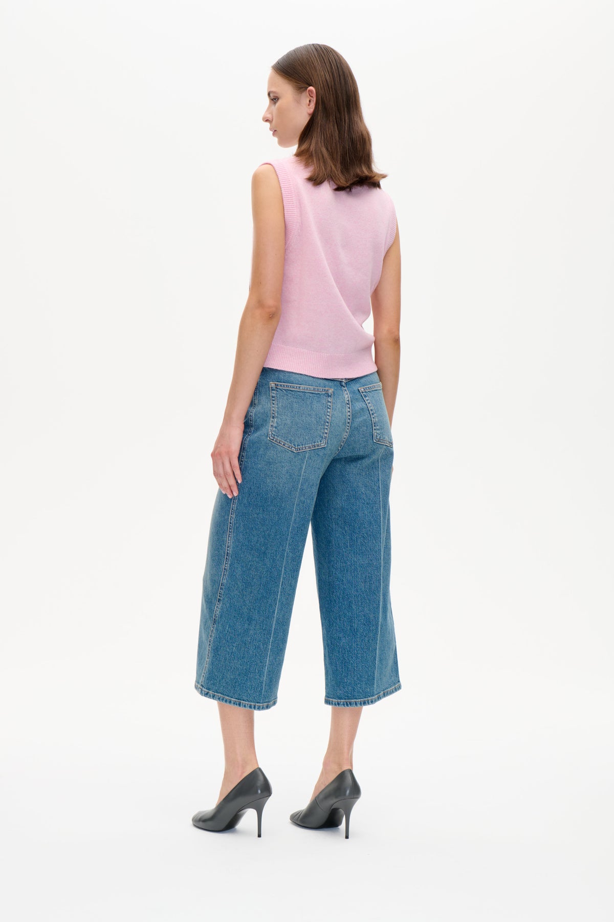 Model trägt blassrosa ärmellose Strickweste und blaue, weit geschnittene, gekürzte Jeans; seitlich und von hinten gezeigt.